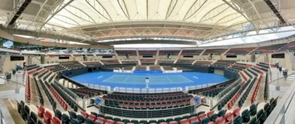 Panoramic view of a modern indoor tennis stadium featuring a blue hard court and empty spectator seating.
