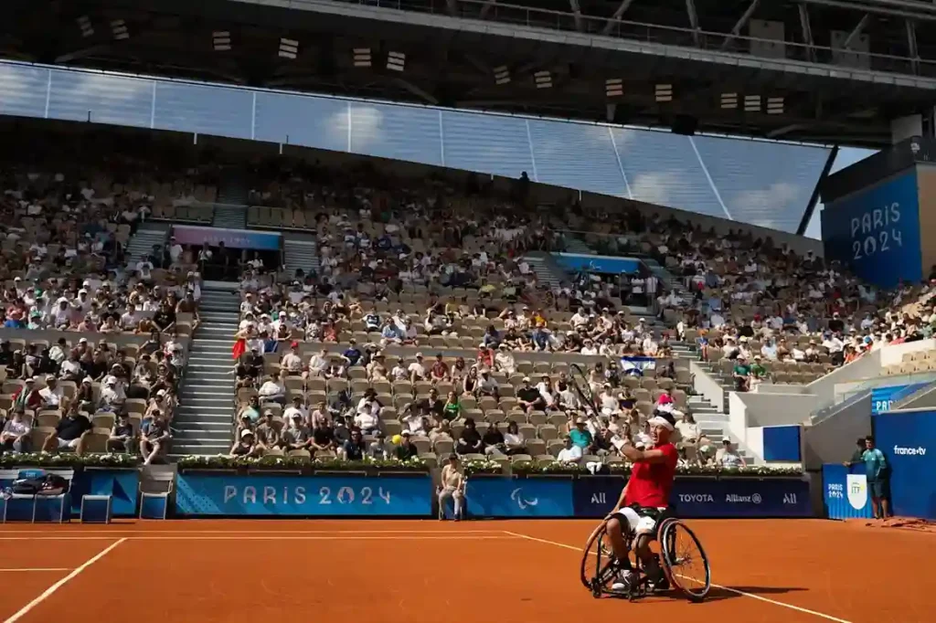 A wheelchair tennis athlete in red shirt preparing a shot on the clay court during the Paris 2024 Paralympics, with a large crowd in the stadium stands.