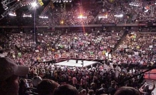 Packed WWE arena with cheering fans surrounding the wrestling ring during a live event.